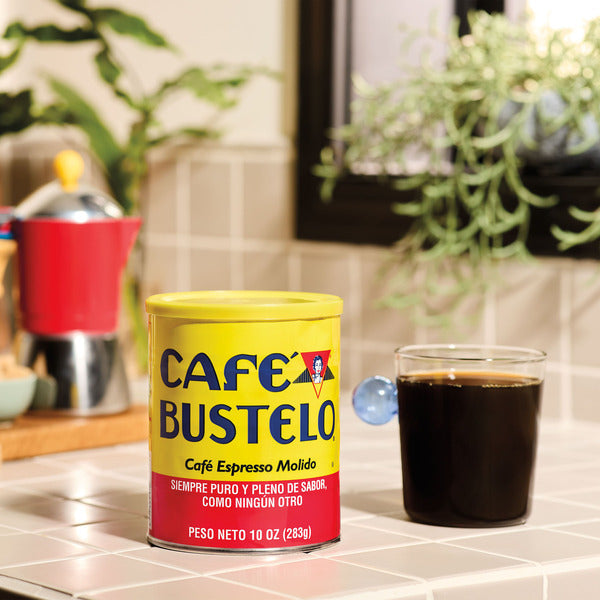 10 oz can of Café Bustelo Espresso Style Dark Roast Ground Coffee beside a freshly brewed glass of black coffee on a bright kitchen countertop.