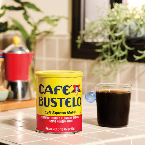 10 oz can of Café Bustelo Espresso Style Dark Roast Ground Coffee beside a freshly brewed glass of black coffee on a bright kitchen countertop.