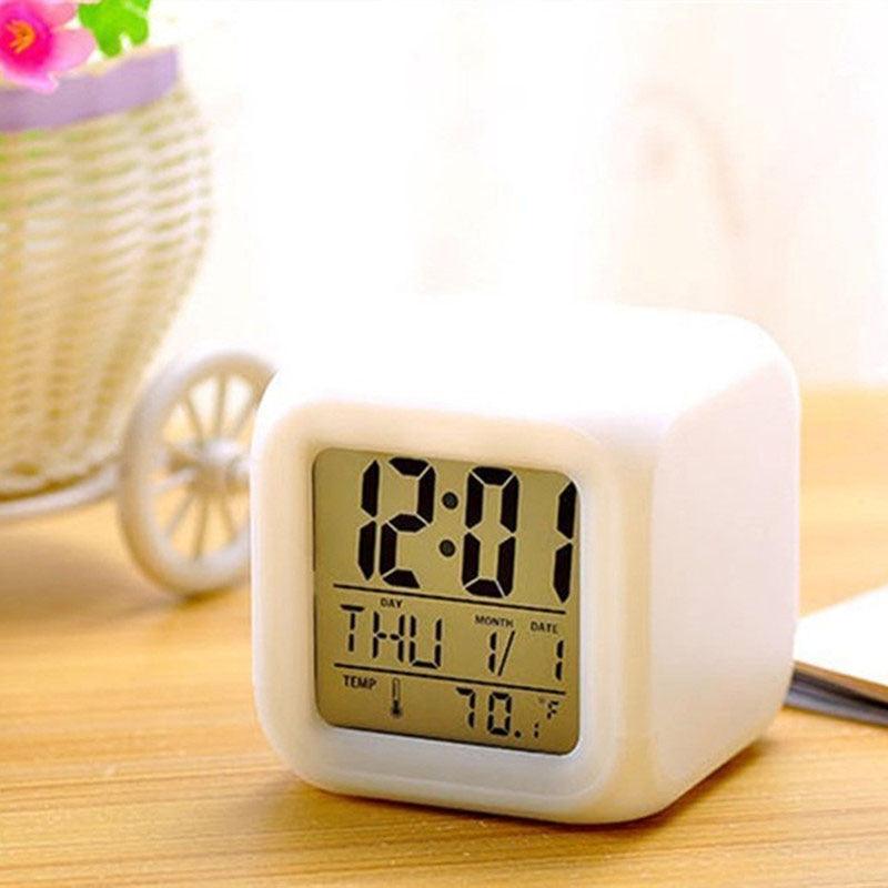 A white cube-shaped digital alarm clock with a yellow-green LCD display showing the time as 12:01 and other settings, placed on a wooden tabletop with a blurred decorative basket in the background.