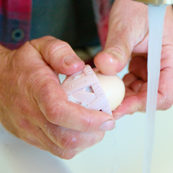 Person using a blush pink GoodEgg silicone egg cleaning brush to gently scrub a fresh egg under running water, demonstrating an easy, hygienic way to clean farm-fresh chicken, duck, goose, and jumbo eggs before use.