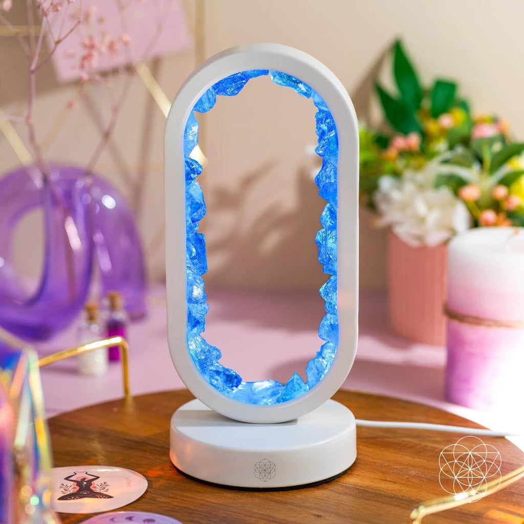Decorative lamp with blue interior on a wooden surface, blurred background with flowers and decor items. Manifest Success - Clear Quartz Lamp of Achievement