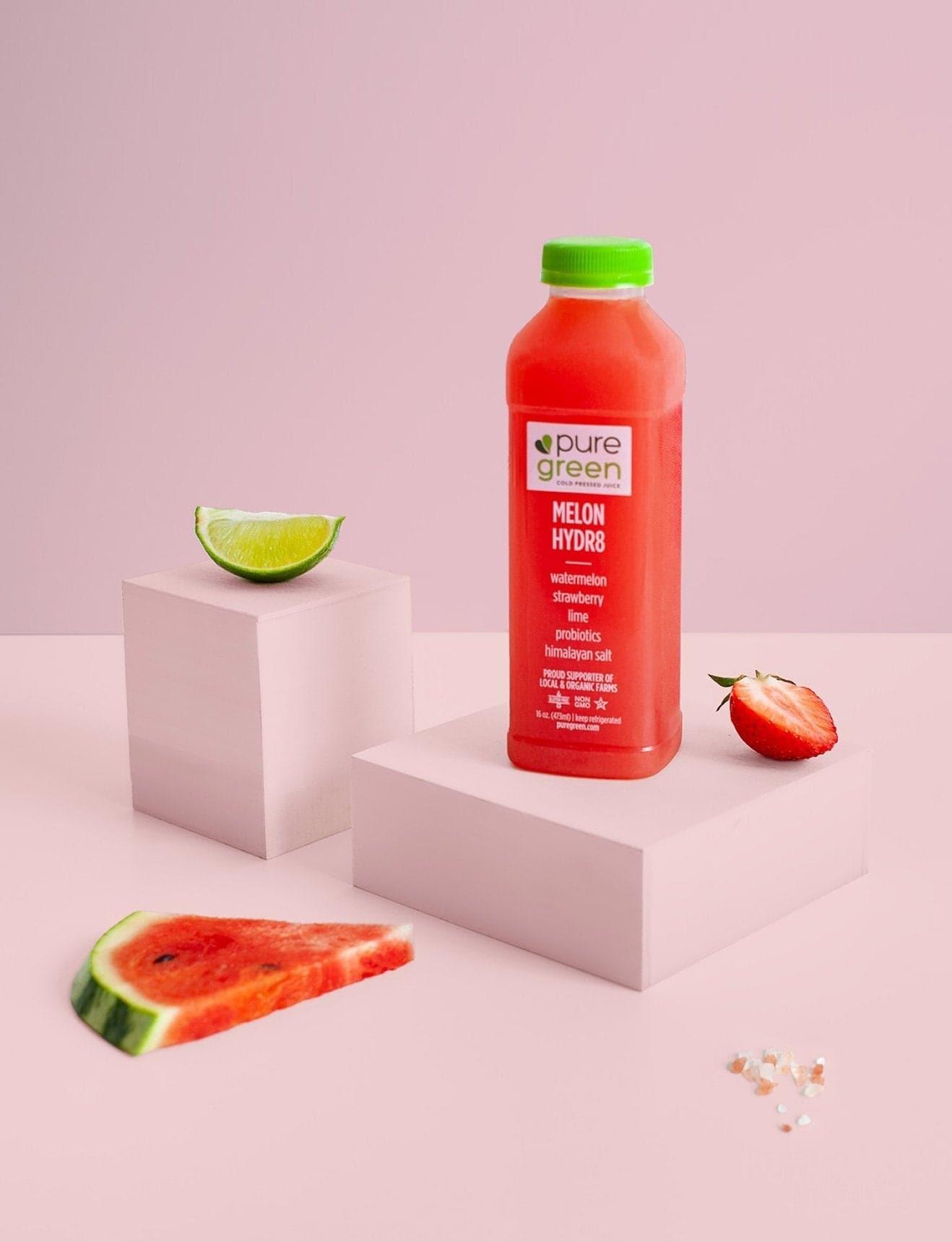 Pure Green juice bottle with watermelon slices and a lime on a pink background. Watermelon, Strawberry, Lime, Probiotics, Himalayan Salt, proud supporters of local and organic farms. The PURE GREEN company cold pressed juice drink.