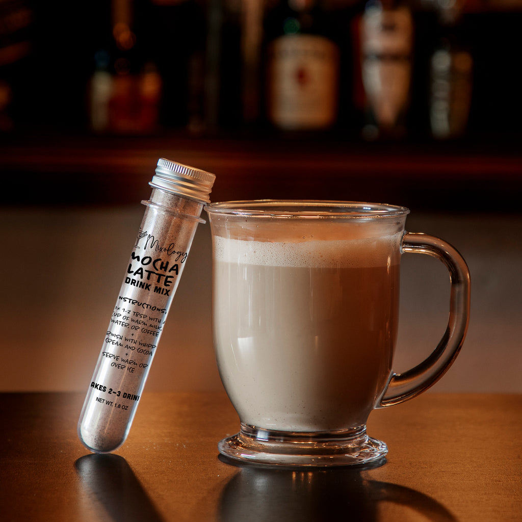Clear glass mug filled with a creamy mocha latte beside a clear vial of Mixology & Co. Mocha Latte Drink Mix on a wooden bar counter, with a warm, softly blurred café background.