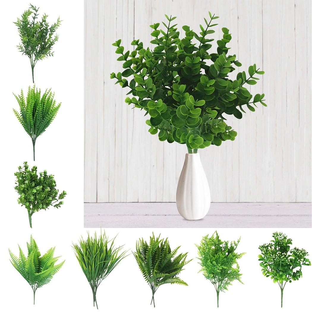 Set of artificial green plants in a white vase against a light wooden panel background.