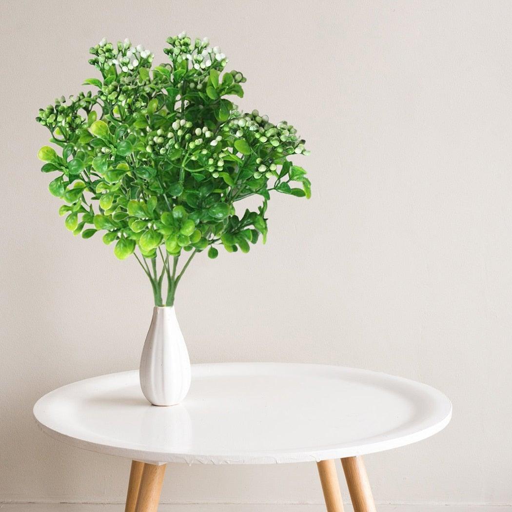 Green artificial plant in a white vase on a round white table with a light gray background