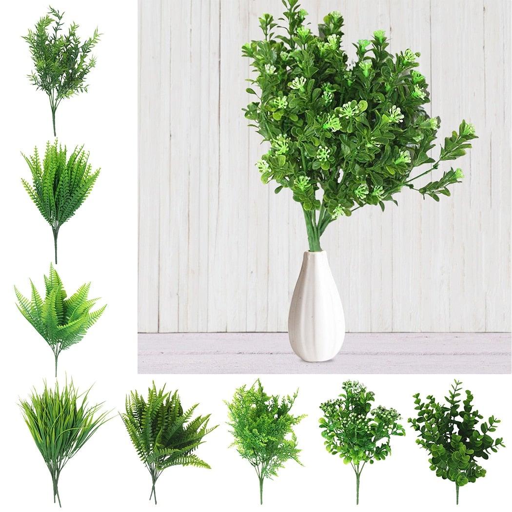 Set of artificial green plants in various styles with a white vase on a light wooden background.