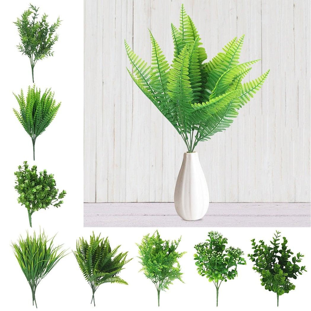 Collection of artificial green plants in various styles with a white vase on a light wooden background.