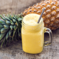 A frothy yellow Passionfruit Pineapple smoothie in a clear glass mason jar with a handle and a black-and-white striped straw, set on a rustic wooden surface with whole pineapples in the background.