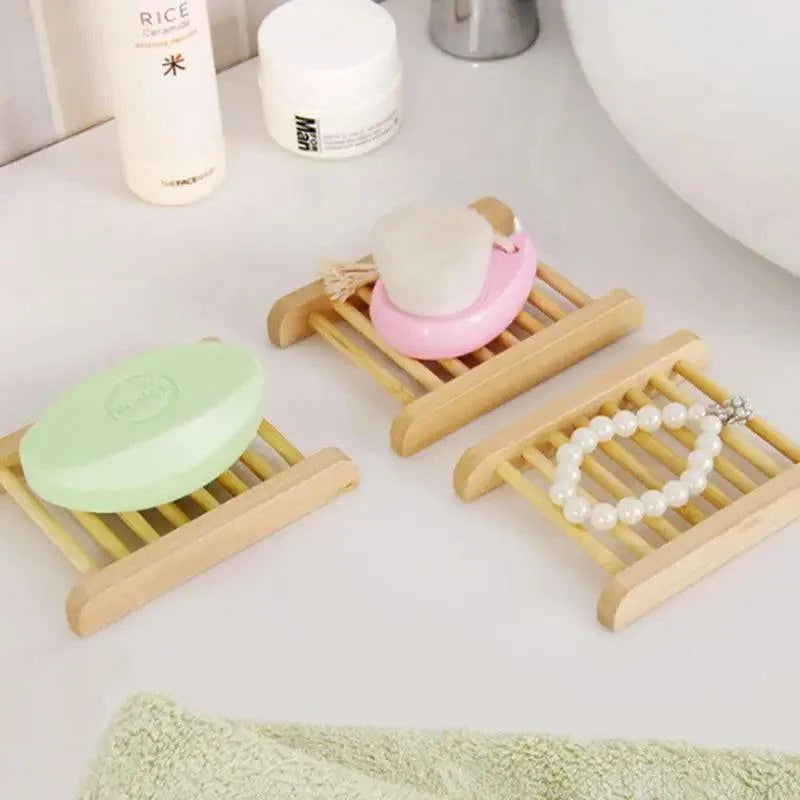 Three light wood slatted soap dish holders arranged on a white bathroom counter, each holding a bar of soap or a pearl bracelet. A mint soap dish with a pale green soap, a pink soap dish with a white soap bar, and a clear soap dish with a white pearl bracelet are shown.