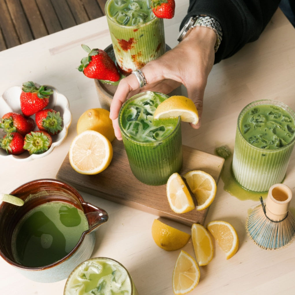 Person holding a glass of green smoothie with strawberries and lemons on a table.