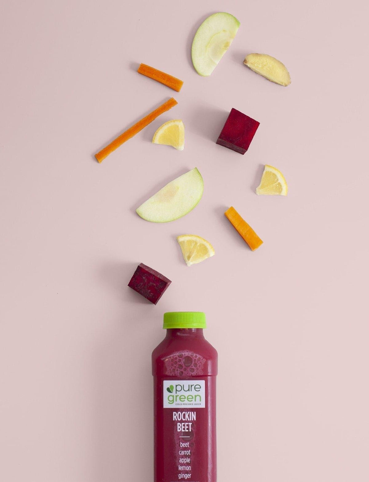 Bottle of 'pure green' juice with beetroot, carrot, and apple pieces on a pink background. Cold-Pressed juice drink with Beets, Carrot, Apple, Lemon and Ginger. A NON-GMO juice drink in 16 - oz bottle. The Pure Green Cold Pressed Juice company is a proud supporter of local and organic farms.