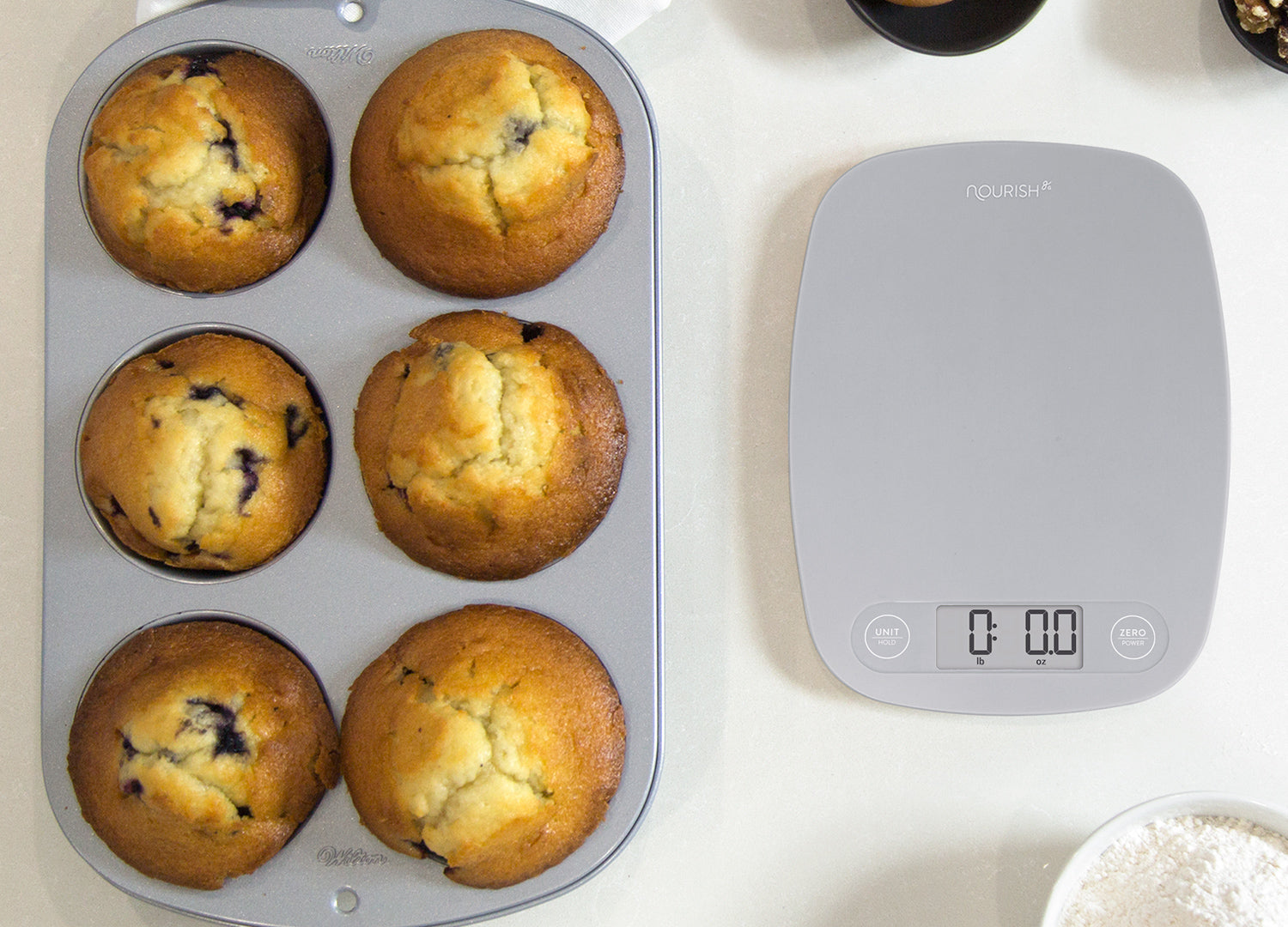 Muffin tray with baked muffins next to a digital kitchen scale on a white surface.