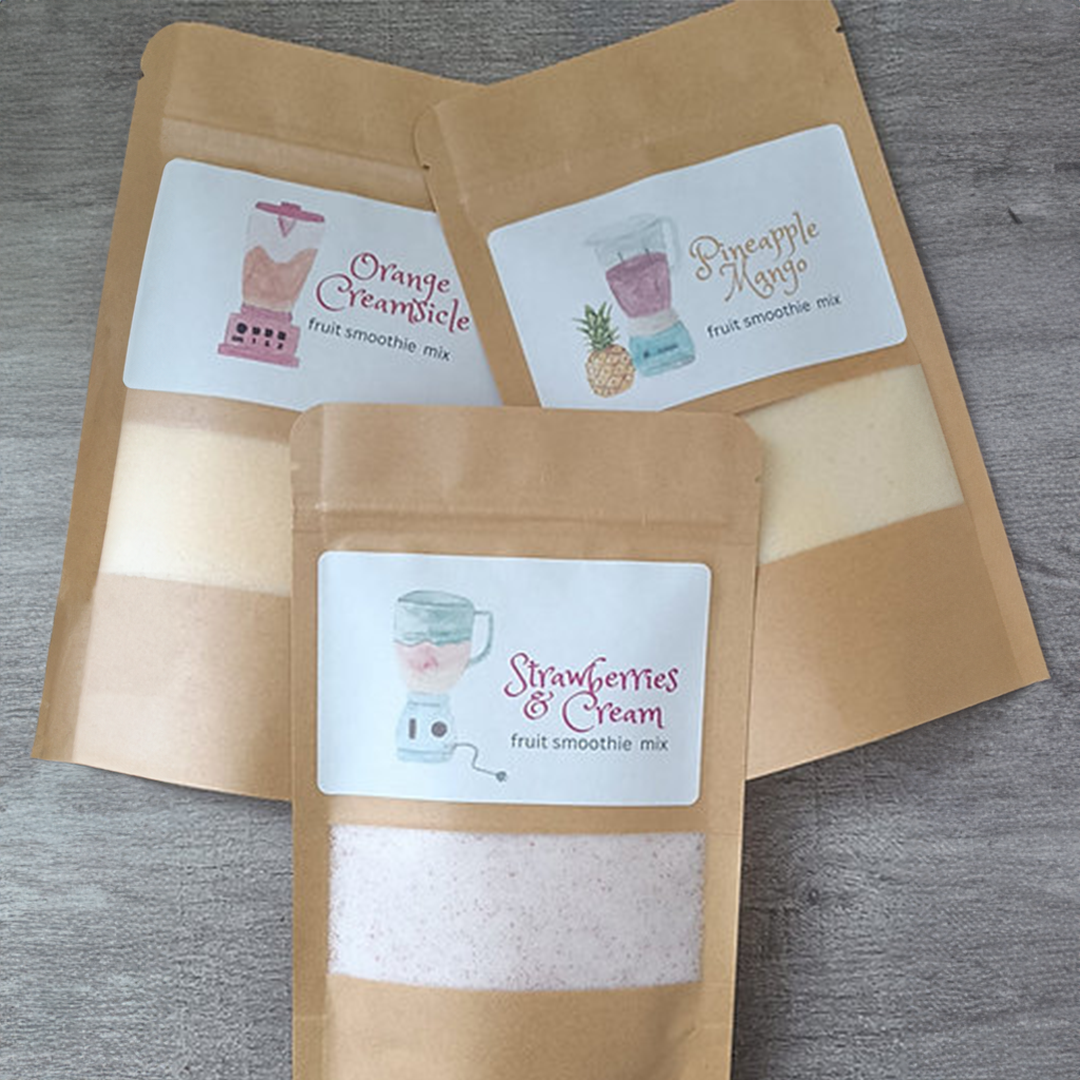 Three kraft paper pouches of premium fruit smoothie mix displayed on a gray wood surface. The front pouch is labeled Strawberries & Cream with a clear window showing pale pink powder, while the two pouches behind feature Orange Creamsicle and Pineapple Mangoflavors, each with colorful blender-themed labels.