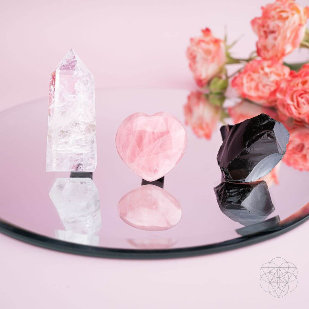 Crystal objects on a reflective surface with pink flowers in the background. Spiritual GPS Guardian Car Vent Crystal Clip Kit | Protection Crystals for Safe Travel & Positive Energy