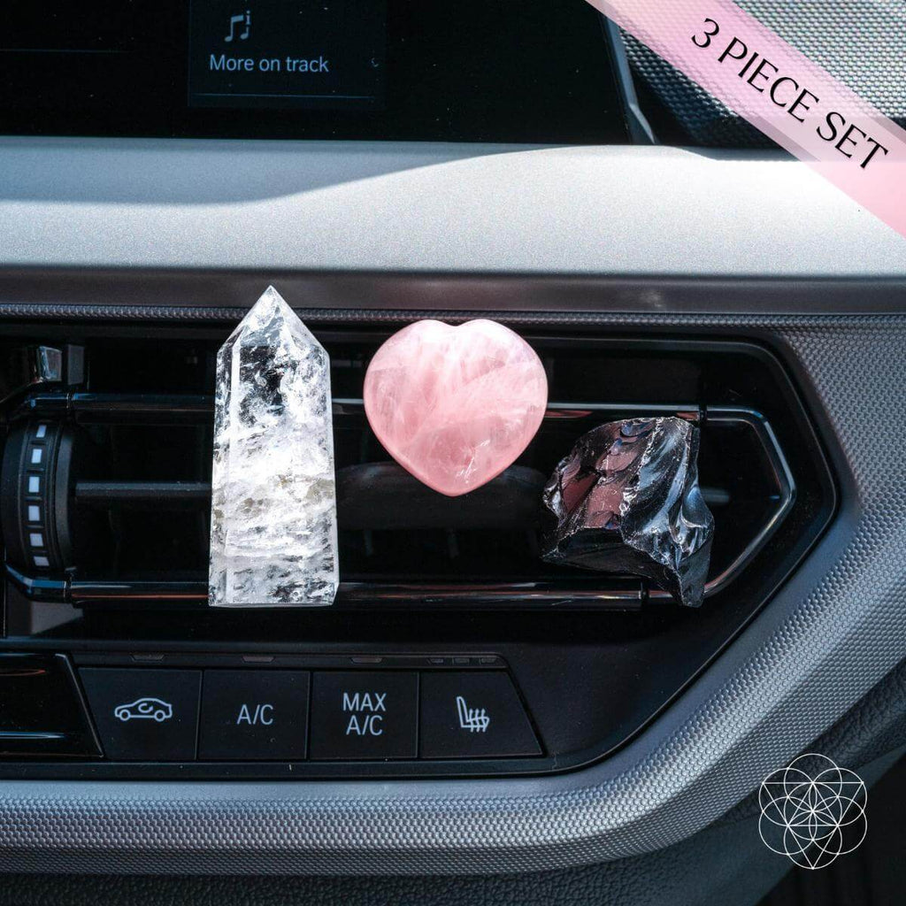 Three crystal objects on a car's air vent with a pink ribbon and text overlay. Car dashboard air vent displaying a 3-piece crystal guardian car kit with a clear quartz point, rose quartz heart, and black obsidian stone, featuring a "3 Piece Set" ribbon overlay. vent clips