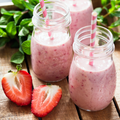 Two clear glass mason jars filled with pink strawberry smoothies, each with a pink-and-white striped straw, set on a wooden surface with fresh strawberries and mint leaves.