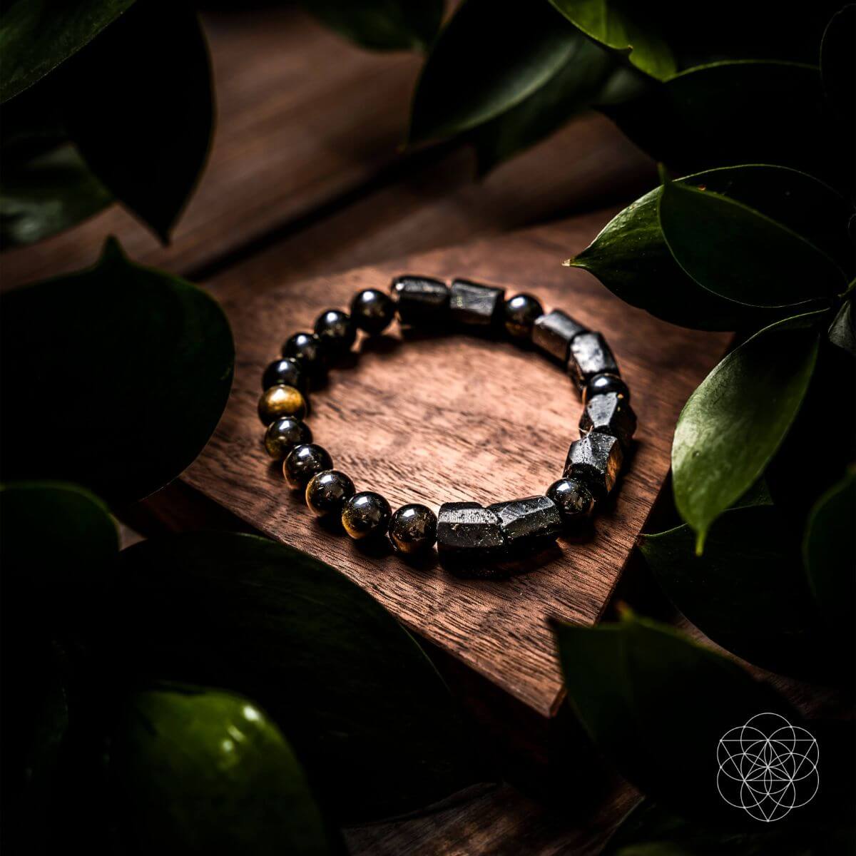A beaded stretch bracelet made of polished black and metallic silver beads with a few gold-toned accents, displayed on a wooden block surrounded by dark green leaves. Direction of Wealth & Success - The Spiritual Protection Bracelet with Black Tourmaline and Golden Obsidian for Absorbing Negativity