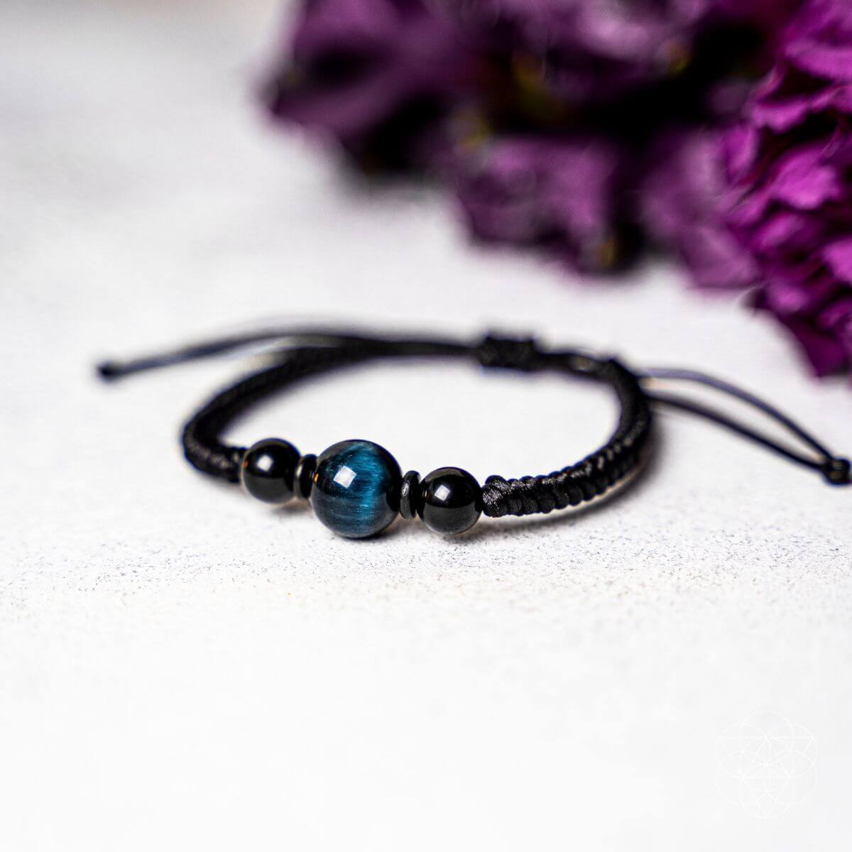 A black beaded bracelet with a central blue-green tiger's eye bead, displayed on a textured light gray surface with blurred purple flowers in the background. The Tiger’s Eye Stability Bracelet: Falcon's Eye (Blue Tiger's Eye) with Black Agate