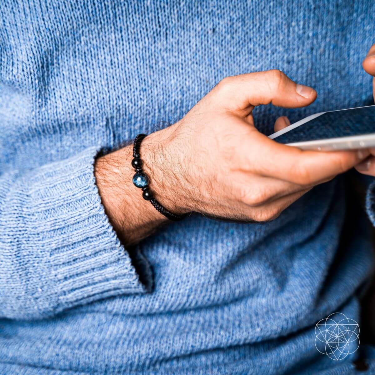 Hand wearing a black beaded bracelet holding a smartphone, with a blue sweater background. The Tiger’s Eye Stability Bracelet: Falcon's Eye (Blue Tiger's Eye) with Black Agate