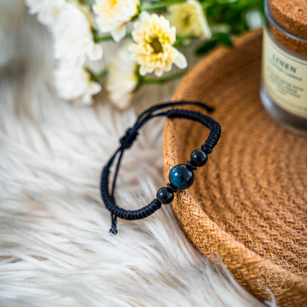 Black beaded bracelet on a fur surface with flowers and a jar in the background. The Tiger’s Eye Stability Bracelet: Falcon's Eye (Blue Tiger's Eye) with Black Agate
