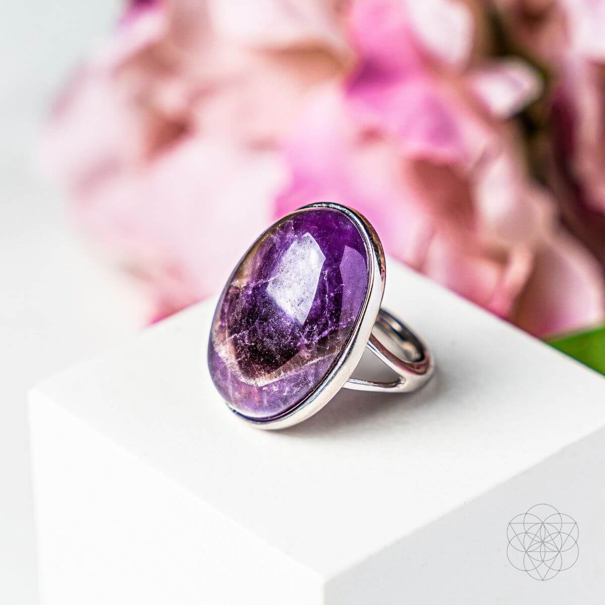 A silver ring featuring a large oval cabochon of purple amethyst with natural white quartz points, displayed on a white block with a soft-focus pink floral background.