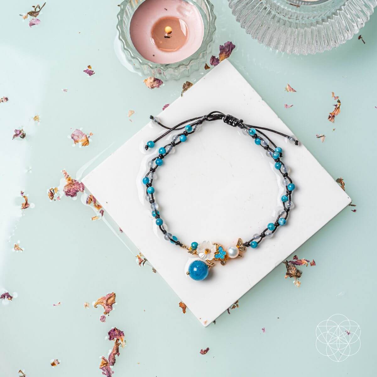 Necklace with blue and black beads on a white square with a candle and decorative elements on a light blue background. Triple Crystal Anklet of Positivity: Pearl, Moonstone, and Apatite