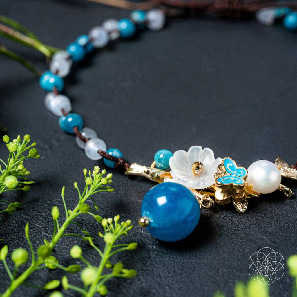 Necklace with blue and white beads and floral elements on a dark surface with green leaves. Triple Crystal Anklet of Positivity: Pearl, Moonstone, and Apatite