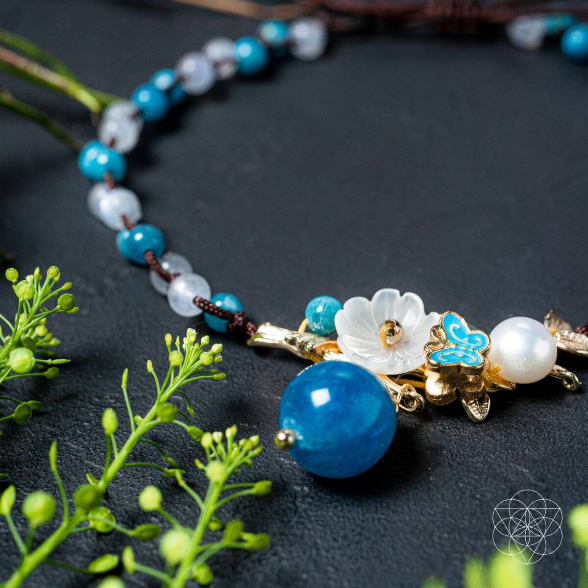 Necklace with blue and white beads and floral elements on a dark surface with green leaves. Triple Crystal Anklet of Positivity: Pearl, Moonstone, and Apatite