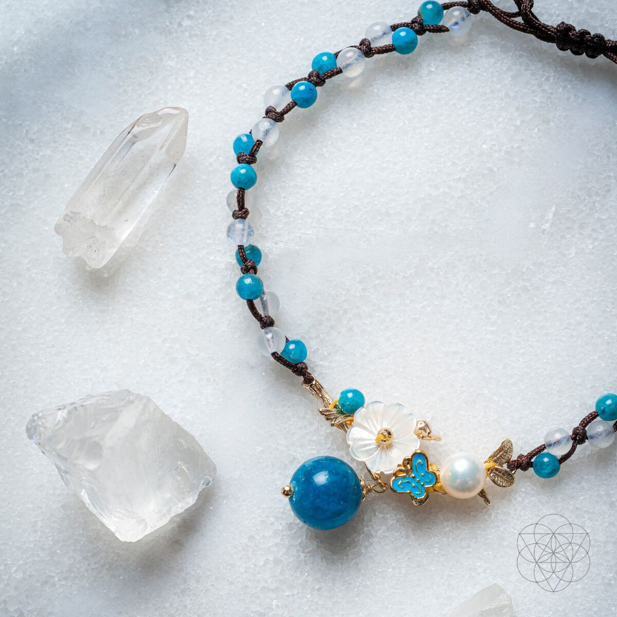 Necklace with blue and white beads on a textured surface with crystals. Triple Crystal Anklet of Positivity: Pearl, Moonstone, and Apatite