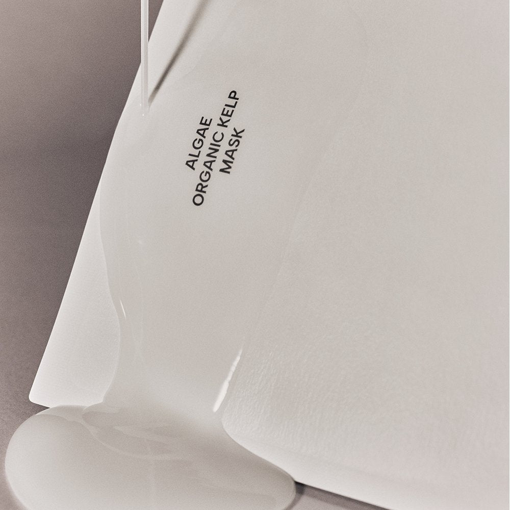 A close-up of a white sheet mask with 'ALGAE ORGANIC KELP MASK' printed in black text, with a glossy, thick white gel being poured over it.