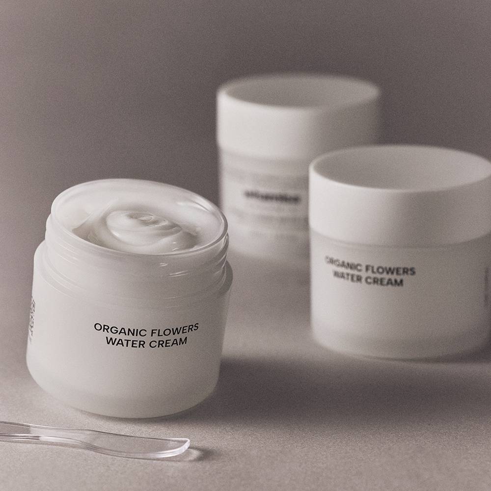 Three white cosmetic jars labeled 'ORGANIC FLOWERS WATER CREAM' on a neutral gray surface, with one jar open in the foreground showing white cream inside and a clear spatula in front.